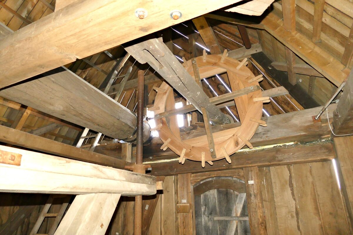 Das große hölzerne Kammrad und die Welle im Dachstuhl der Bockwindmühle, von Lichtstrahlen durchzogen.